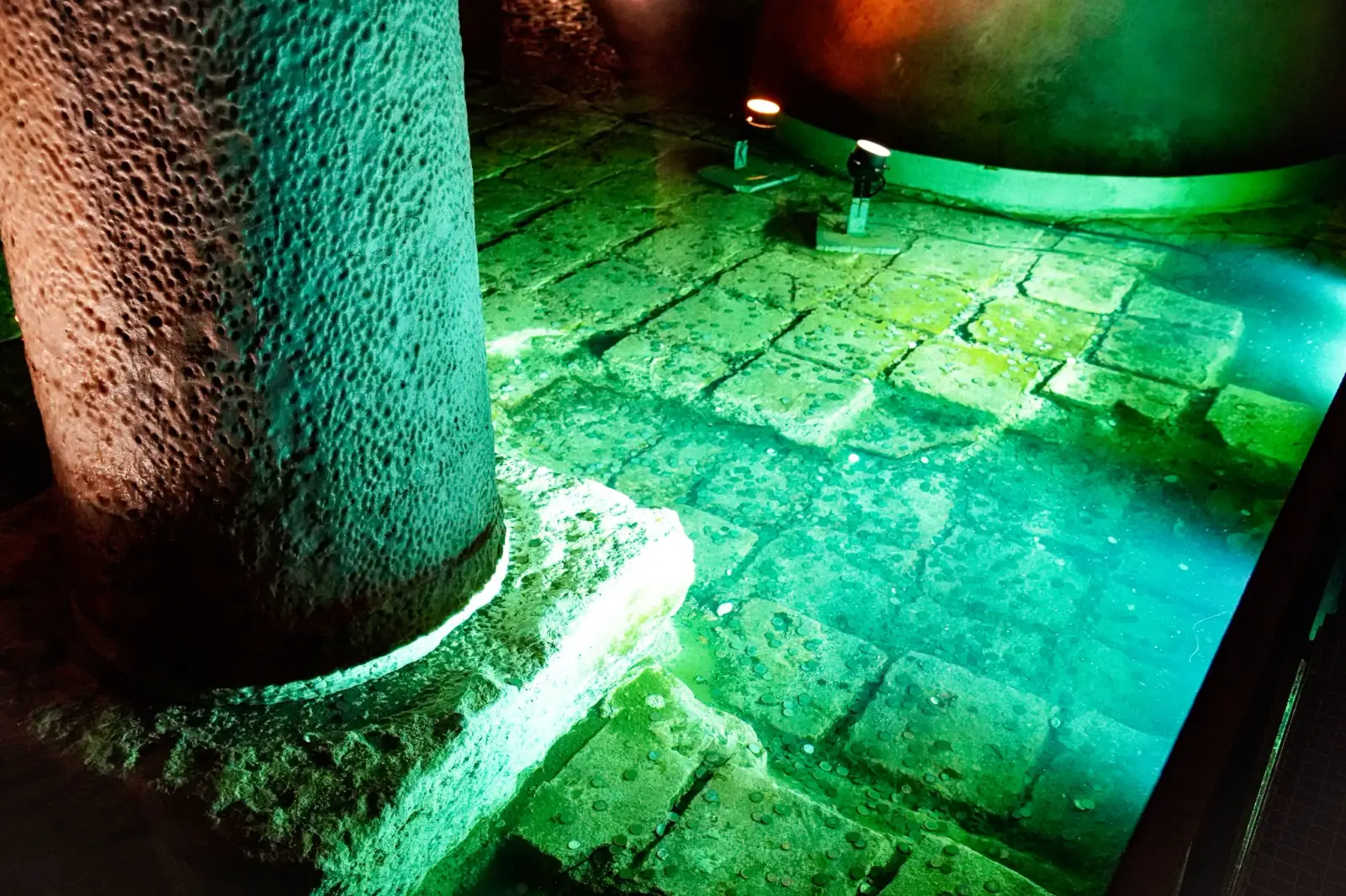 The foot of a column and the brick-paved floor beneath the shallow water, lit green.