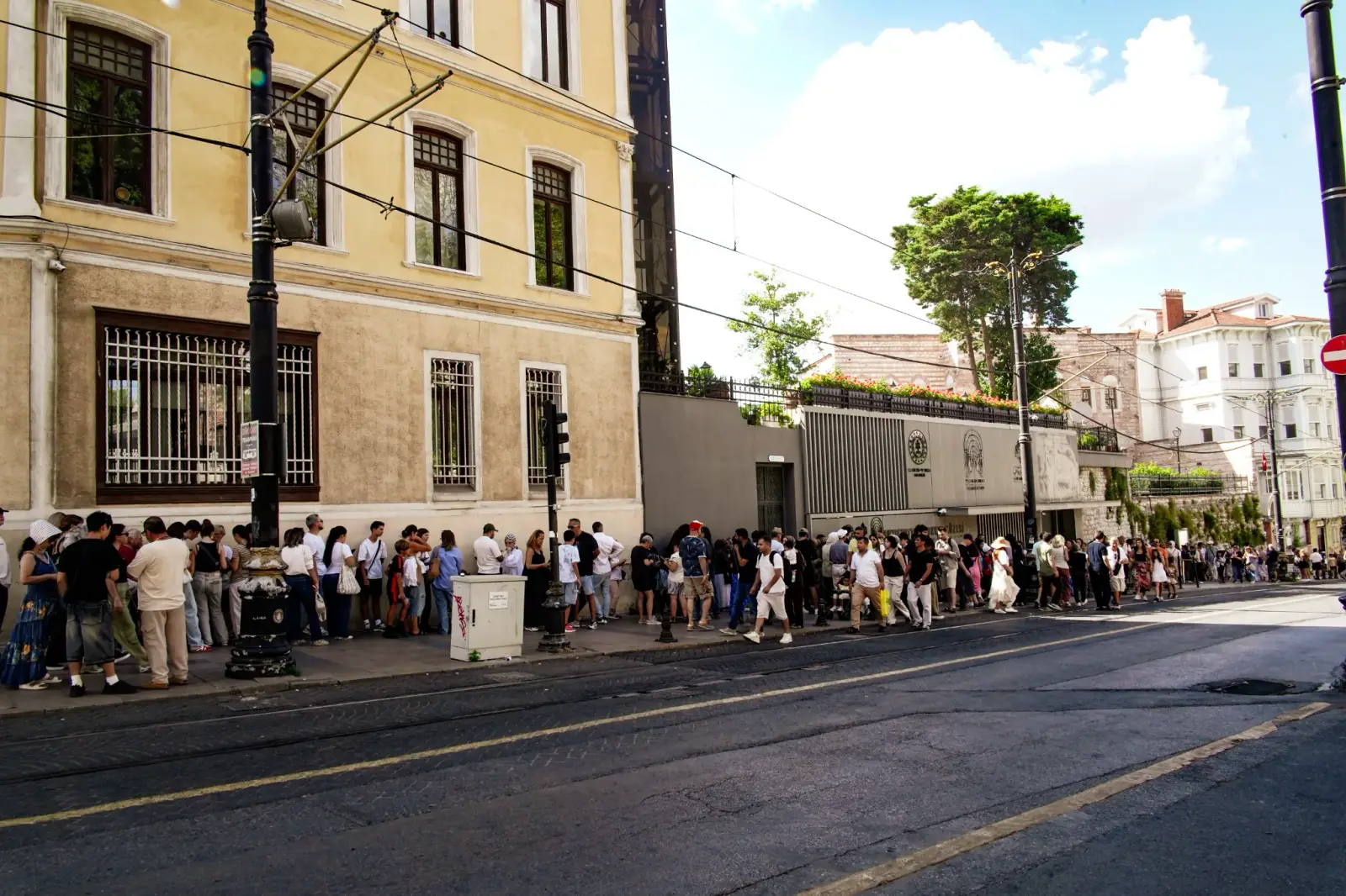 The entrance seen along the street, tram rails and overhead wires in the foreground and the queue running past the wall.