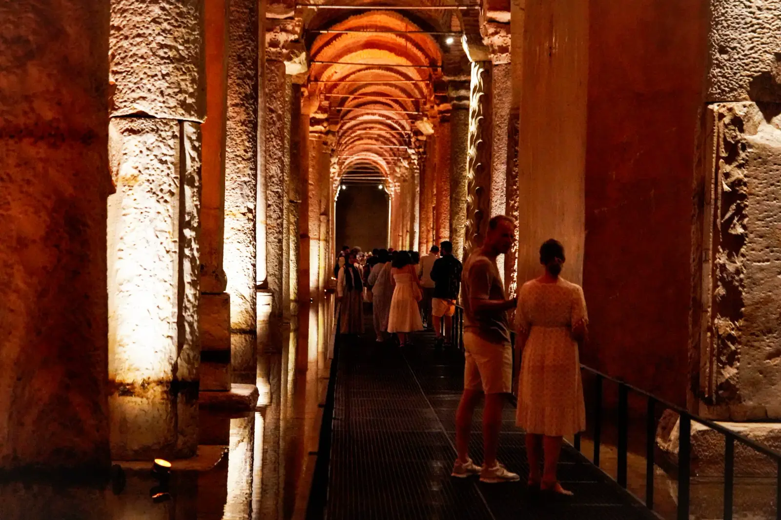 A lit corridor between two rows of columns, brick arches receding into the dark, visitors on the walkway.